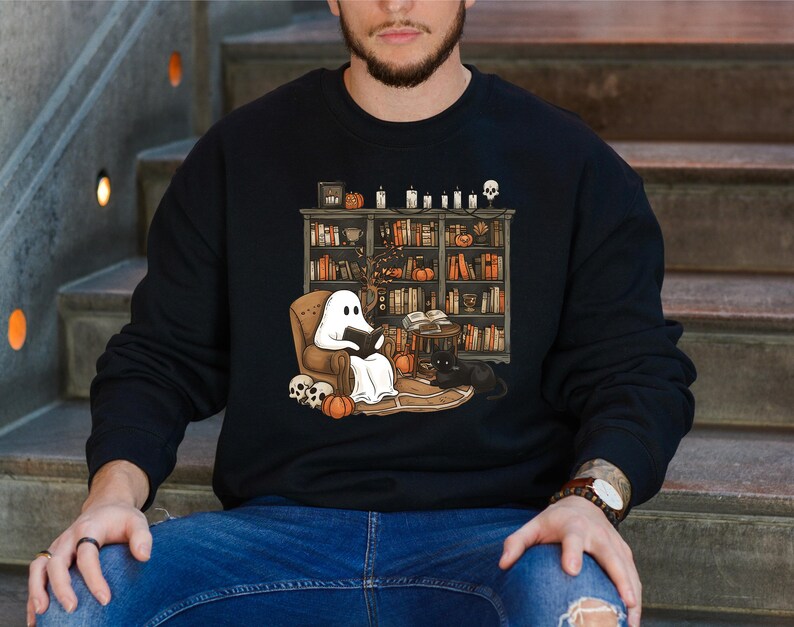 May include: Black sweatshirt featuring a Halloween-themed illustration. A ghost reads in a cozy armchair in front of a bookshelf filled with books, pumpkins, and candles. A black cat sits nearby. Skulls and pumpkins are also present.
