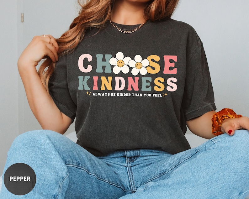 Peut inclure: T-shirt gris fonc&eacute; avec l'inscription "CHOOSE KINDNESS" dans un style r&eacute;tro color&eacute;. Le motif comprend des marguerites souriantes et le texte "ALWAYS BE KINDER THAN YOU FEEL".