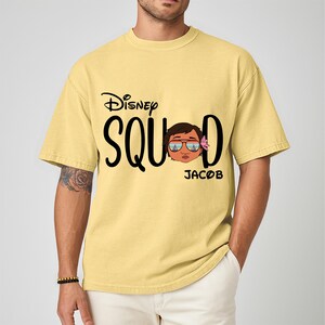 May include: A light yellow t-shirt with the words "Disney Squad" in black script. The letter "O" in "Squad" is replaced with a cartoon face wearing sunglasses and a flower. The name "Jacob" is printed below the face.