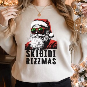 Puede incluir: Una sudadera beige con un gr&aacute;fico de Pap&aacute; Noel con gafas de sol y un gorro rojo. El texto "Skibidi Rizzmas" est&aacute; impreso debajo del gr&aacute;fico.