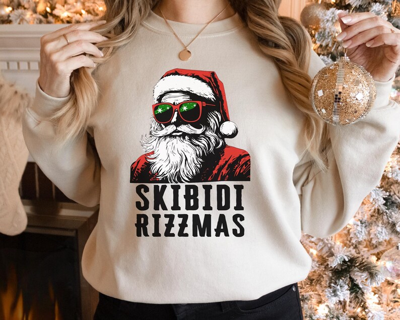 Puede incluir: Una sudadera beige con un gr&aacute;fico de Pap&aacute; Noel con gafas de sol y un gorro rojo. El texto "Skibidi Rizzmas" est&aacute; impreso debajo del gr&aacute;fico.