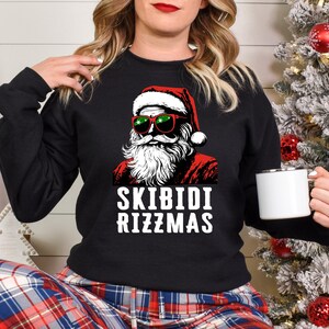 Puede incluir: Sudadera negra con un gr&aacute;fico de Santa Claus con gafas de sol y un gorro rojo. El texto "Skibidi Rizzmas" est&aacute; impreso debajo del gr&aacute;fico.