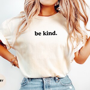 Op de afbeelding: Een wit T-shirt met de tekst "be kind." in zwart gedrukt op de voorkant. Het shirt wordt gedragen door een persoon met lang blond haar. Het shirt is ivoor van kleur.