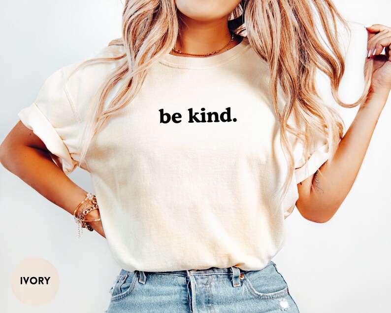 Op de afbeelding: Een wit T-shirt met de tekst "be kind." in zwart gedrukt op de voorkant. Het shirt wordt gedragen door een persoon met lang blond haar. Het shirt is ivoor van kleur.