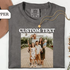 May include: A gray Comfort Colors t-shirt with a family photo and the words "CUSTOM TEXT" in white. A gold star pendant necklace is draped across the shirt. A brown hat is visible in the upper left corner.