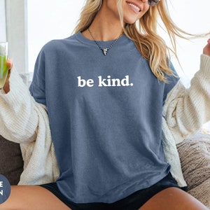 Op de afbeelding: Een vrouw draagt een oversized t-shirt van blauwe jeans met de tekst "be kind." in wit.