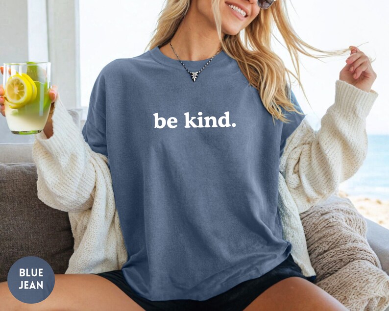 Op de afbeelding: Een vrouw draagt een oversized t-shirt van blauwe jeans met de tekst "be kind." in wit.