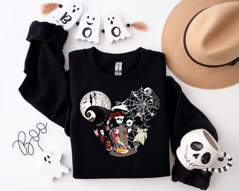 Puede incluir: Sudadera negra con dise&ntilde;o en forma de coraz&oacute;n con personajes de "Pesadilla antes de Navidad" y una luna llena. Incluye una telara&ntilde;a blanca, fantasmas y una taza con forma de calavera. Tambi&eacute;n se ve un sombrero color canela y la palabra "Boo".