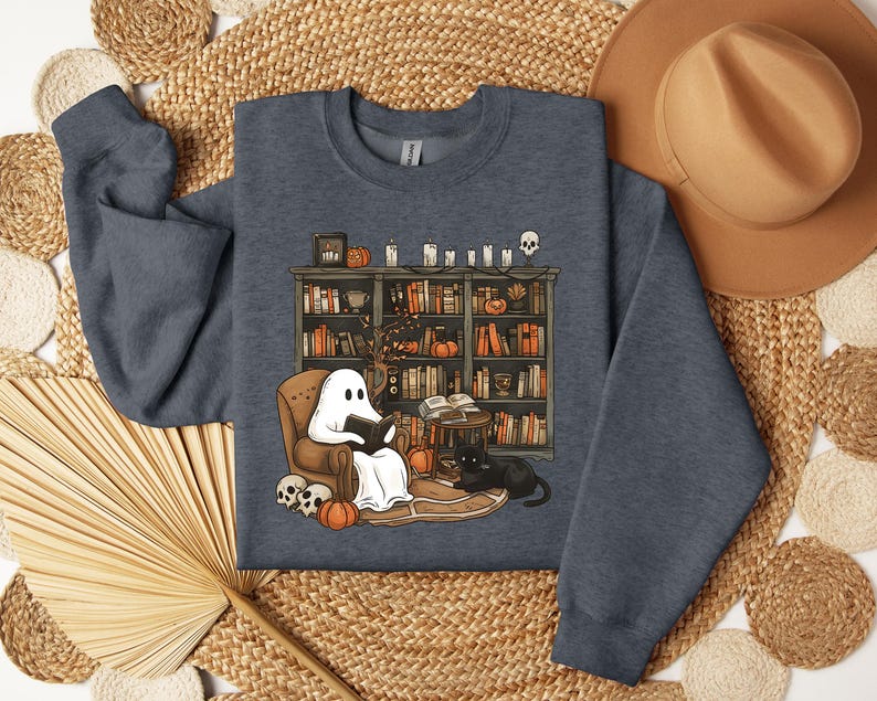 May include: A dark gray sweatshirt featuring a Halloween-themed illustration. The design depicts a ghost reading in a cozy armchair in front of a bookshelf, with pumpkins, candles, and a black cat. Skulls and fall decor complete the scene.