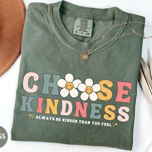 Peut inclure: Un t-shirt vert mousse avec l'inscription "CHOOSE KINDNESS" dans une police r&eacute;tro, avec des graphiques de marguerites avec des visages souriants. La phrase "ALWAYS BE KINDER THAN YOU FEEL" est en dessous. Un collier en or est pos&eacute; sur le t-shirt.