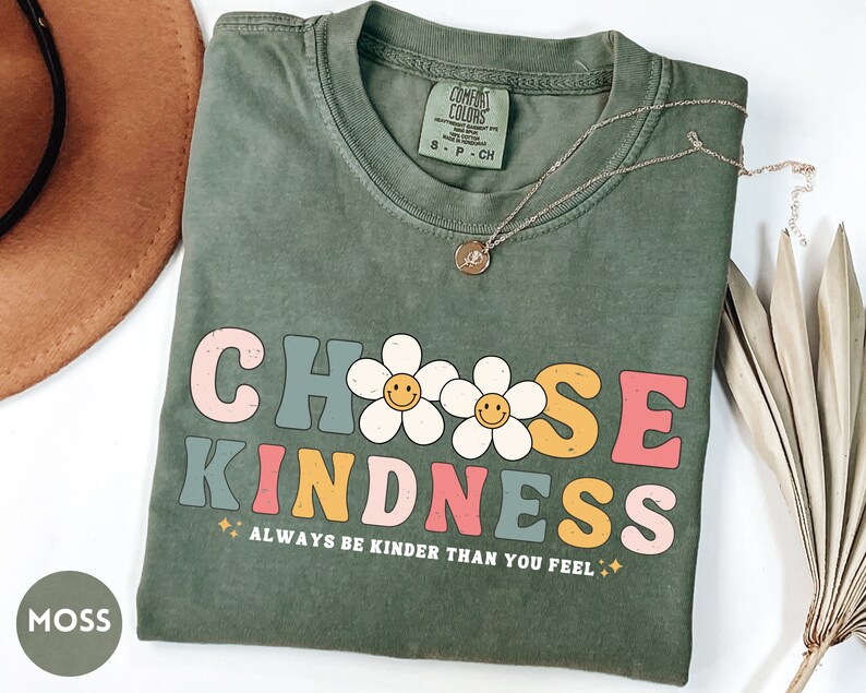 Peut inclure: Un t-shirt vert mousse avec l'inscription "CHOOSE KINDNESS" dans une police r&eacute;tro, avec des graphiques de marguerites avec des visages souriants. La phrase "ALWAYS BE KINDER THAN YOU FEEL" est en dessous. Un collier en or est pos&eacute; sur le t-shirt.