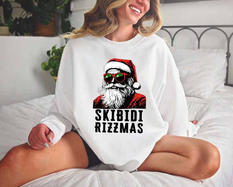 Puede incluir: Sudadera blanca con un gr&aacute;fico de Pap&aacute; Noel con gafas de sol y un gorro rojo. El texto "Skibidi Rizzmas" est&aacute; impreso debajo del gr&aacute;fico.