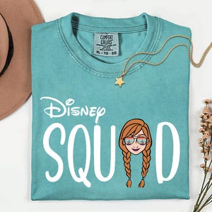 K&ouml;nnte beinhalten: T&uuml;rkisfarbenes Comfort Colors T-Shirt mit dem Text "Disney SQUAD" und einem Cartoon-Bild einer Figur mit Sonnenbrille. Eine goldene Sternanh&auml;nger-Halskette und ein brauner Hut sind ebenfalls im Bild.