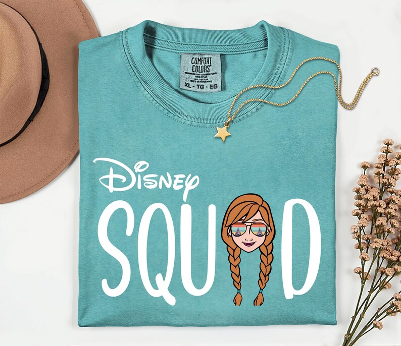 K&ouml;nnte beinhalten: T&uuml;rkisfarbenes Comfort Colors T-Shirt mit dem Text "Disney SQUAD" und einem Cartoon-Bild einer Figur mit Sonnenbrille. Eine goldene Sternanh&auml;nger-Halskette und ein brauner Hut sind ebenfalls im Bild.