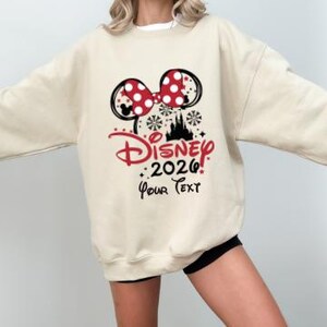 Może przedstawiać: Kremowa bluza z motywem Minnie Mouse. Projekt zawiera czerwoną kokardę w kropki, czarne uszy myszy, słowo "Disney" w czerwonym skrypcie, rok "2026" i tekst "Your Text."