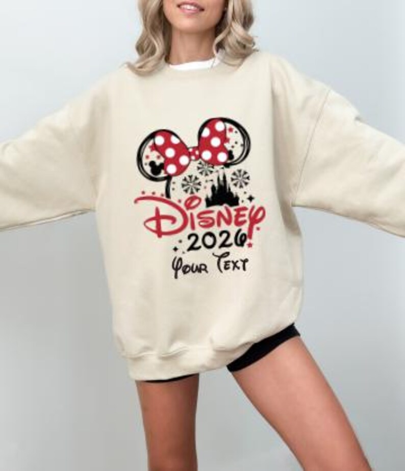 Może przedstawiać: Kremowa bluza z motywem Minnie Mouse. Projekt zawiera czerwoną kokardę w kropki, czarne uszy myszy, słowo "Disney" w czerwonym skrypcie, rok "2026" i tekst "Your Text."
