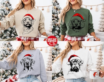 May include: A collection of sweatshirts featuring dog portraits wearing Santa hats. The sweatshirts are in various colors, including beige, green, gray, and white. Each sweatshirt has a different dog breed design with a name below the dog's portrait. The text "NEXT DAY SHIPPING" is also visible.