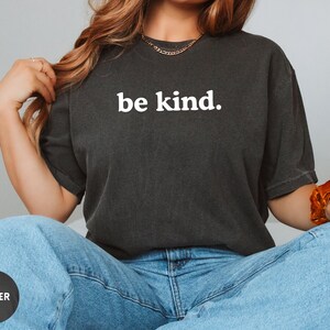 Op de afbeelding: Een zwart T-shirt met de tekst "be kind." in wit gedrukt op de voorkant. Het shirt wordt gedragen door een persoon met lang bruin haar.
