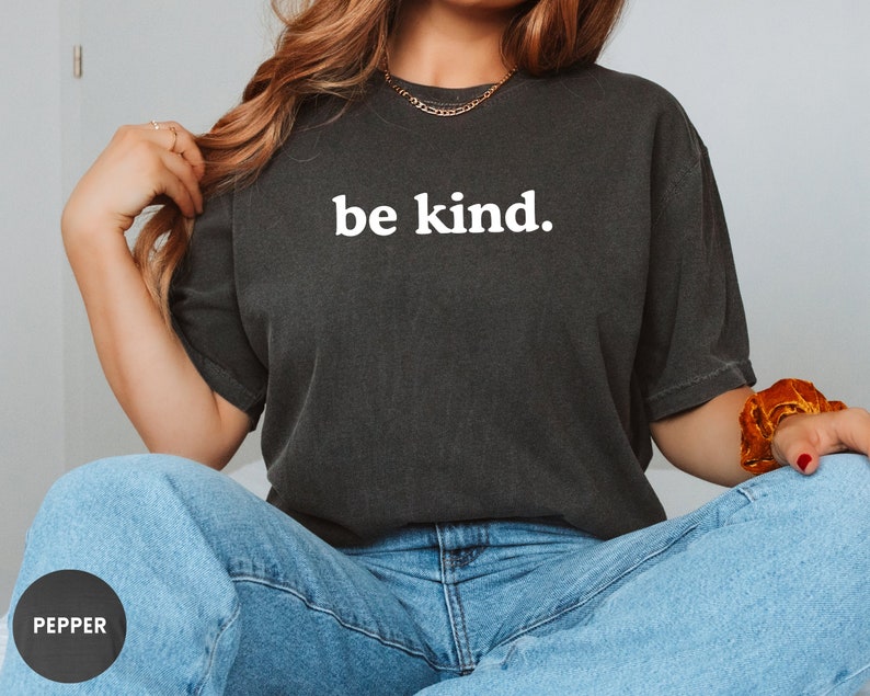 Op de afbeelding: Een zwart T-shirt met de tekst "be kind." in wit gedrukt op de voorkant. Het shirt wordt gedragen door een persoon met lang bruin haar.
