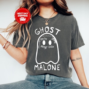 May include: Dark grey t-shirt featuring a white ghost design with the text "GHOST MALONE". The ghost has two oval eyes and the words "Always Tired" below. The shirt is a comfortable, casual style.