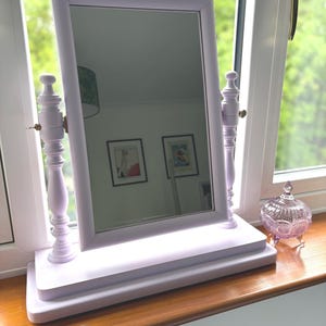 May include: A lilac-painted vanity mirror with a rectangular frame and decorative supports. The mirror is on a matching base, and a small glass jar is next to it. The mirror reflects a room with framed pictures.