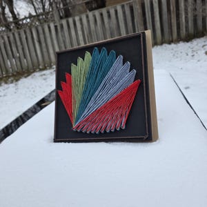 May include: A square string art piece with a fan design, using red, green, teal, and grey threads on a black background. The artwork is framed in a brown wooden frame, creating a striking contrast. A handmade craft item.