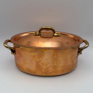 May include: A round copper pot with a matching lid and two side handles. The pot features a polished metallic finish with warm orange and brown tones. The lid has a central handle, and both the lid and side handles are made of brass.