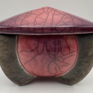 May include: A ceramic lidded vessel featuring a deep magenta and purple crackle glaze on the lid and central body, contrasted by matte dark gray or charcoal-colored sides. The container has a rounded, organic shape with a wide, flat-topped lid.
