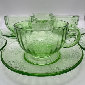 Può includere: Un set di tazze da tè e piattini in vetro verde. Le tazze e i piattini presentano un motivo a uva e foglie. Le tazze hanno manici e poggiano su piattini abbinati. Il set è realizzato in vetro verde trasparente.