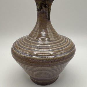 May include: A ceramic vase with a wide, rounded body and a narrow neck. The surface features horizontal, concentric ridges throughout the body, finished in a mottled brown and grey glaze with a darker, textured rim at the top opening.