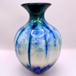 Może przedstawiać: Ceramiczny wazon z krystalicznym szkliwem, o pękatym korpusie i rozszerzanym wylocie. Powierzchnia posiada gradient od głębokiego błękitu u góry i u podstawy, przechodzący w jaśniejszy błękit nieba i biel, z wyraźnymi kolistymi formacjami kryształów.