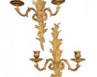 May include: A pair of gold-toned metal wall sconces, each featuring two candle holders extending from a central vertical stem. The design includes ornate leaf-like foliage and a decorative ribbon bow detail at the centre of each sconce.