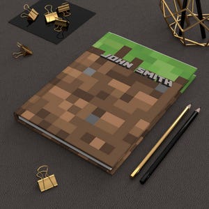 May include: A brown and green notebook with a Minecraft-inspired design. The cover features a pixelated image of a dirt block with grass on top. The name "JOHN SMITH" is printed in white on the cover.