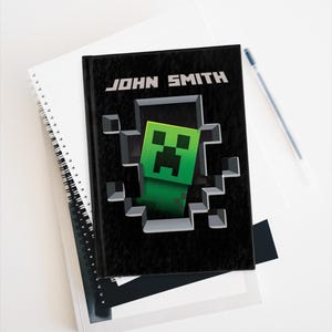 May include: Black notebook featuring a Minecraft Creeper character emerging from a pixelated wall. The notebook is personalised with the name "JOHN SMITH" printed on the front.