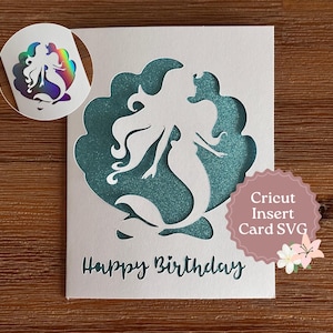 May include: A white card with a mermaid silhouette cut-out design, revealing a teal glittery background. The words "Happy Birthday" are printed at the bottom. A circular sticker with a holographic mermaid is in the upper left corner.