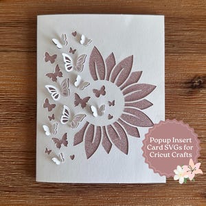 Può includere: Un biglietto bianco con un design ritagliato di un girasole e farfalle. Il girasole è di colore oro rosa glitterato e le farfalle sono bianche e oro rosa. Il biglietto ha il testo "Popup Insert Card SVGs for Cricut Crafts".