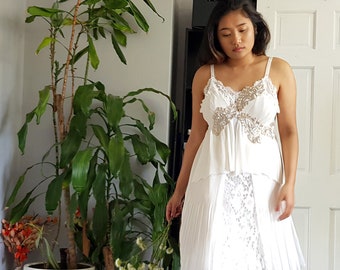 May include: A woman wearing a white lace dress with a ruffled top and a pleated skirt. She is standing in a room with a mirror and plants.