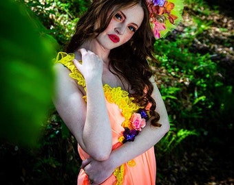 May include: A woman wearing a peach dress with yellow lace trim and a floral crown. The crown is made of green tulle and has pink, purple, and orange flowers.