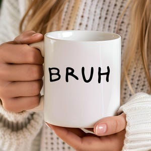 K&ouml;nnte beinhalten: Eine wei&szlig;e Keramiktasse mit einer schwarzen handschriftlichen Aufschrift, die "BRUH" lautet.