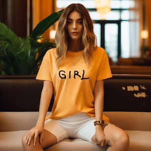 K&ouml;nnte beinhalten: Eine Frau tr&auml;gt ein gelbes T-Shirt mit dem Aufdruck "GIRL". Sie sitzt auf einem Sofa und tr&auml;gt wei&szlig;e Shorts.