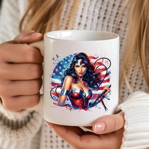 May include: White ceramic mug featuring a vibrant illustration of a superheroine with a patriotic theme. The design includes the American flag, stars, and a woman in a red and blue costume. The mug is held by a person.
