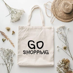 K&ouml;nnte beinhalten: Eine wei&szlig;e Canvas-Tragetasche mit schwarzem Text, der in handschriftlicher Form "GO SHOPPING" lautet. Die Tasche befindet sich auf einer wei&szlig;en Oberfl&auml;che mit getrockneten Blumen und einer geflochtenen Strohhut.