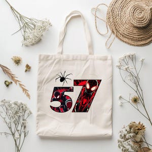 Może przedstawiać: Biała pł&oacute;cienna torba typu tote z czerwoną i czarną grafiką Spider-Mana w numerze 57. Czarny pająk znajduje się w lewym g&oacute;rnym rogu wzoru.