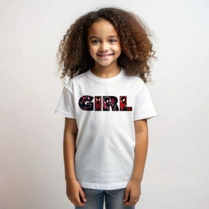 Może przedstawiać: Biała koszulka z nadrukiem graficznym słowa "GIRL" czarnymi literami. Litery są wypełnione czerwono-czarną grafiką superbohatera.