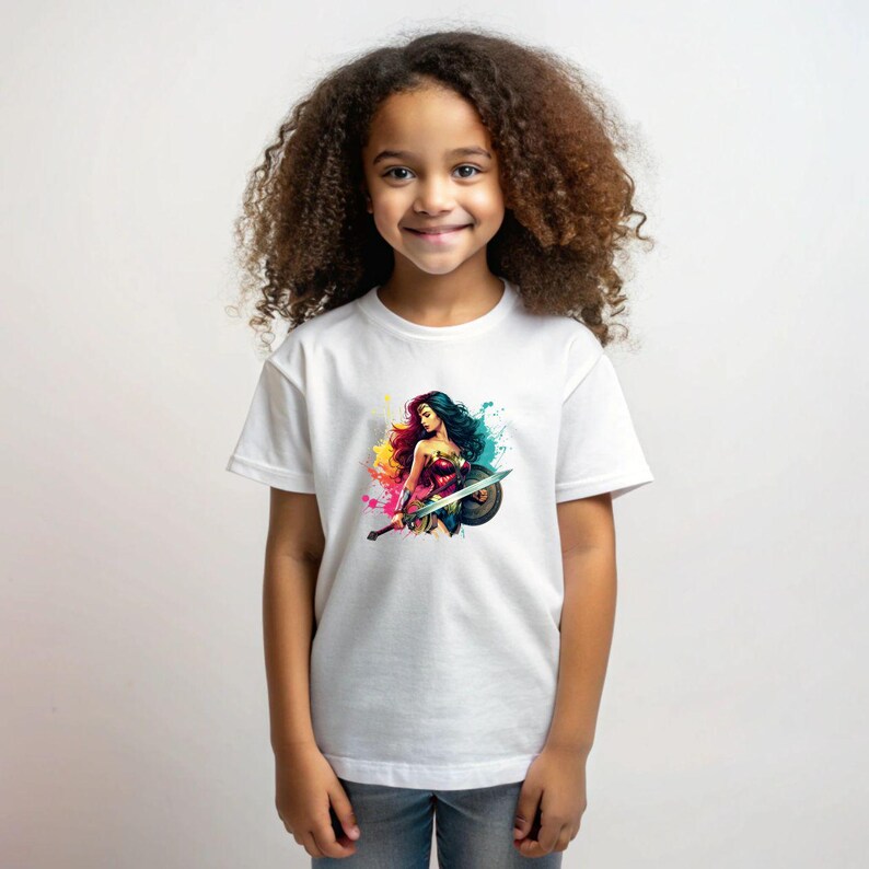 May include: White t-shirt featuring a colorful illustration of Wonder Woman with a sword and shield. The graphic includes splashes of yellow, pink, blue, and green. The shirt is short-sleeved and appears to be made of cotton.