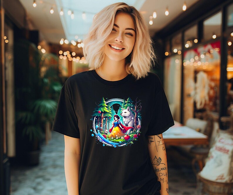 Op de afbeelding: Een zwart T-shirt met een kleurrijke grafische print van Sneeuwwitje en een wolf in een bossetting. De grafiek gebruikt een blauw, groen en paars kleurenschema.