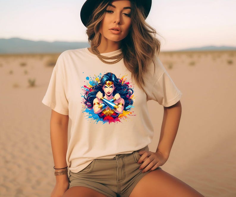 May include: A white t-shirt with a colorful graphic of Wonder Woman with her arms crossed. The graphic is surrounded by colorful paint splatters.