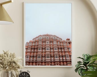 May include: A framed print of a detailed, coral-colored building with numerous windows and a domed roof. The building is set against a pale sky. The print is displayed on a wooden shelf with a clock, vase, and plant.