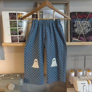 May include: Blue and grey chequered trousers featuring an elastic waistband and two white ghost appliqués on the front legs. The trousers are hung on a wooden hanger, showcasing a repeating square pattern across the fabric and simple ghost designs.