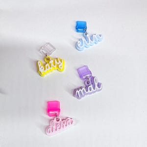 Puede incluir: Cuatro pendientes de nombre acrílicos de colores en amarillo, azul, morado y rosa. Cada pendiente presenta un nombre diferente en una fuente cursiva. Los pendientes están sujetos a un pequeño cierre cuadrado.